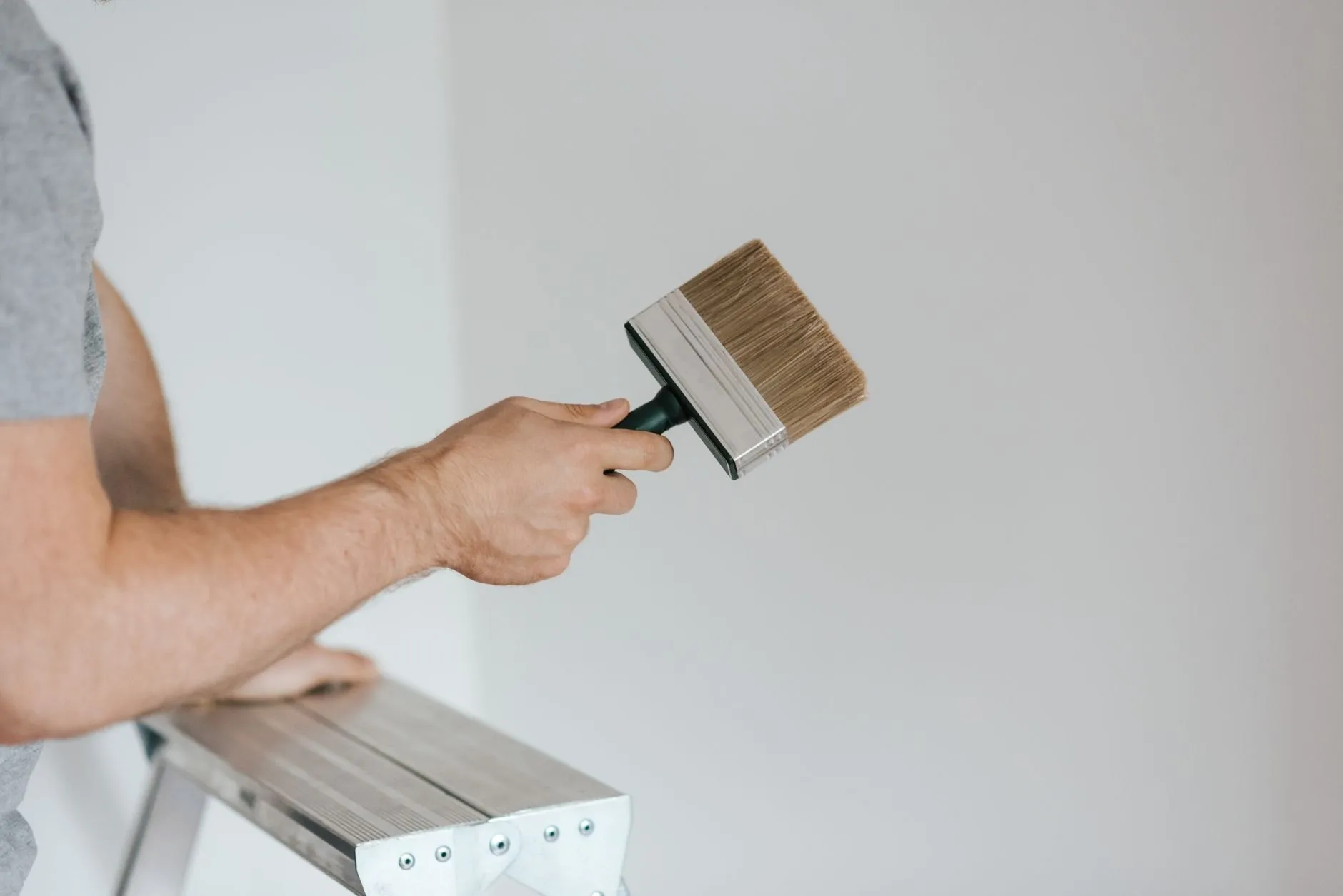 A hand holding a wide paintbrush up to a pale wall beside a stepladder