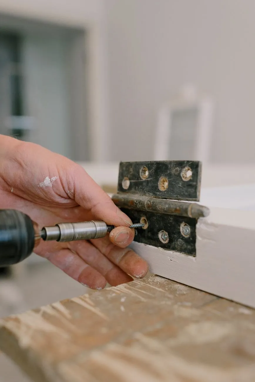 A hand holding a screw into a black butt hinge on a white door edge while a driver turns it