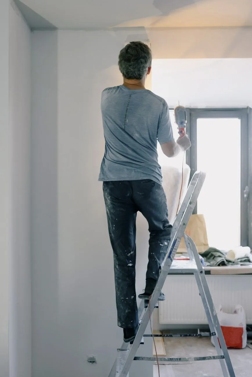 A handyman on a stepladder working near the ceiling of a bright, half-decorated room