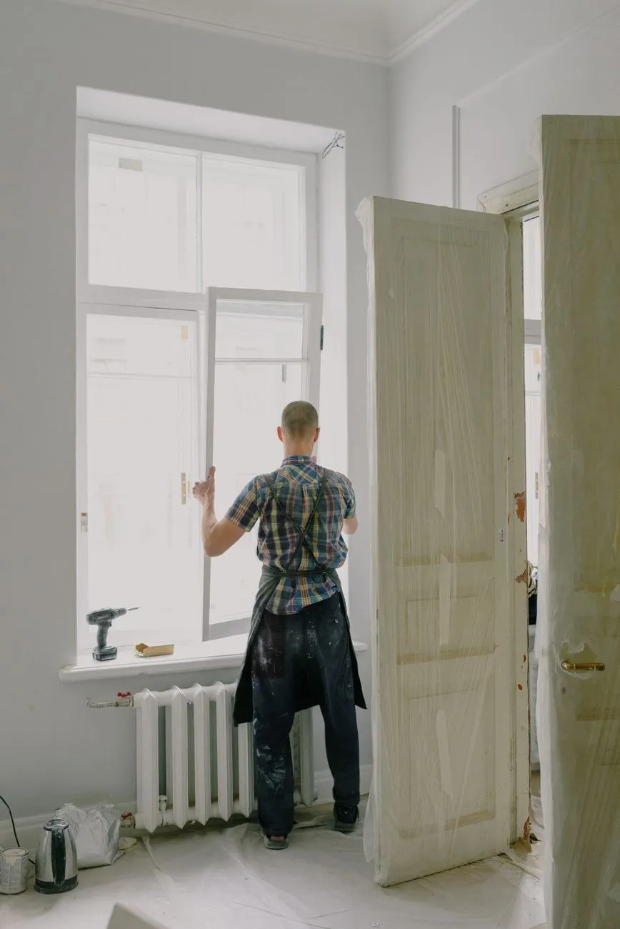 A handyman fitting a tall sash window in a bright room, the inner door open beside him