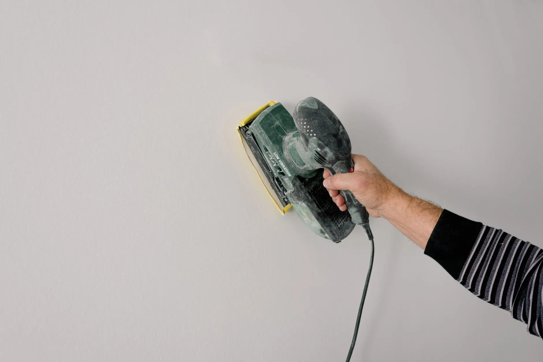 A hand holding an orbital sander flat against a filled and smoothed wall
